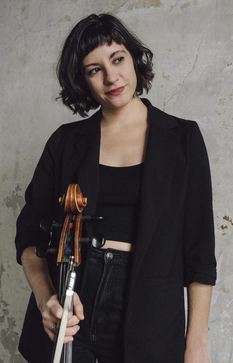 A photo of Iva Casian Lakos, a person with long black hair and dark clothes. Iva is holding a stringed instrument.